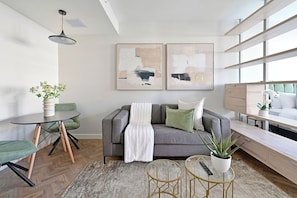 A modern living room with a grey sofa, a round coffee table, and a small side table. There are abstract paintings on the wall, a potted plant, and a window with a view. - Stylish 1-Bed Apart | Iconic High-Rise City Views (Cape Town)