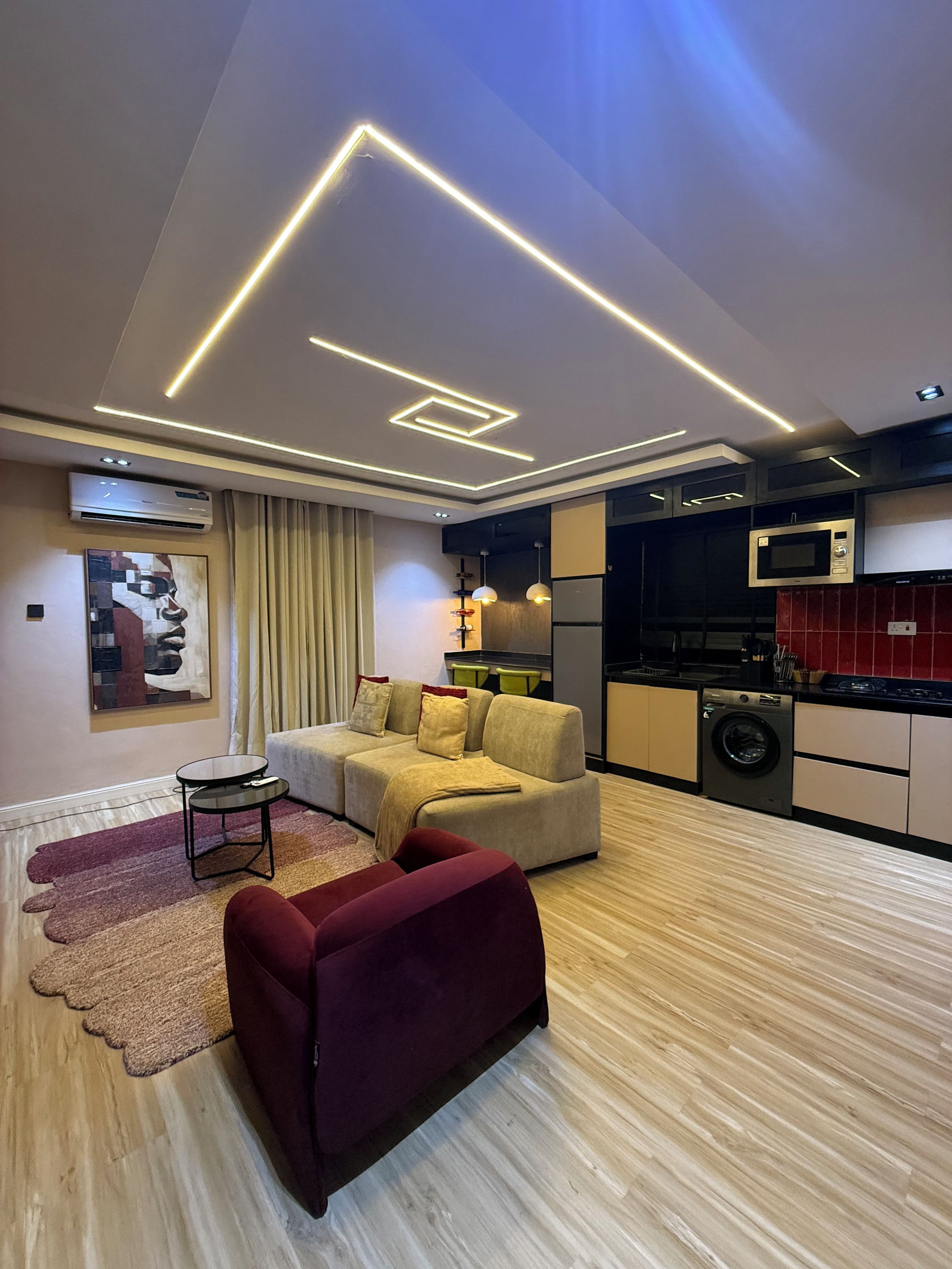 Un salon moderne avec un canapé d’angle, une petite table ronde et un fauteuil rouge. La pièce est dotée d’un plancher en bois et d’un plafond agrémenté de luminaires aux formes géométriques.