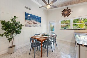 A dining area with a wooden table set for four, a ceiling fan, a large painting of a beach scene, and a window with a view of greenery. - The Coco Plum, Hot Tub, Island Living (West Palm Beach)