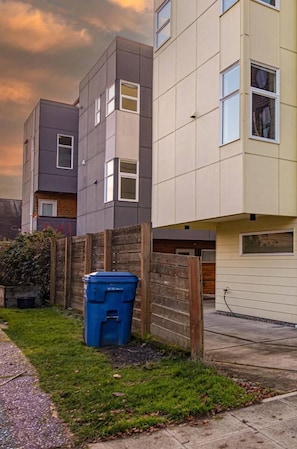 A modern two-story residential building with a flat roof, a wooden fence, and a blue trash bin in the yard. - Spacious Modern House Rooftop Backyard Loft 3B2.5B (Seattle)