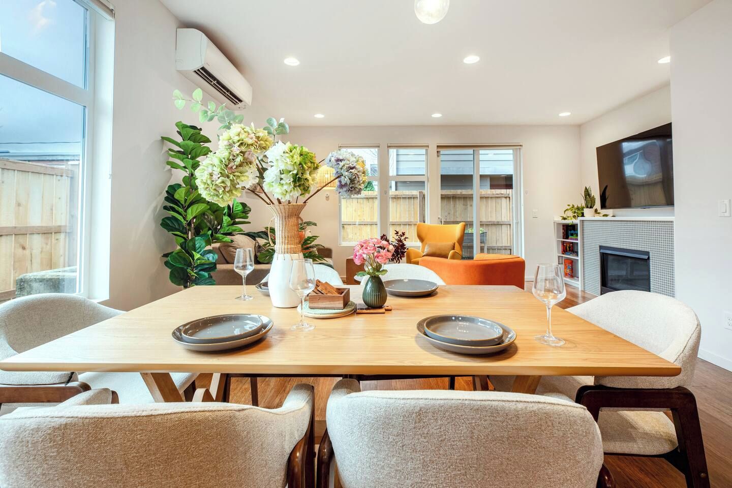 A dining area with a wooden table set for four, surrounded by chairs, with a vase of flowers and a television in the background.