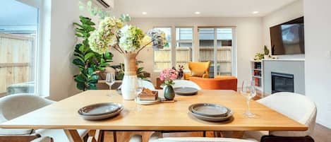 A dining area with a wooden table set for four, surrounded by chairs, with a vase of flowers and a television in the background.