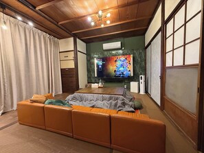 A living room with a sectional sofa, a flat-screen TV, and a ceiling light fixture. - maicca2: A Traditional Japanese House in Numazu, Eura—Entire House Rental | Sleeps Up to 8 · BBQ · 100-inch TV · 1-minute walk to the marina (沼津市)