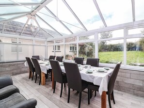 A conservatory with a dining table set for six, surrounded by chairs, and large windows offering a view of a garden. - Herriotts Bridge Cottage (Bristol)