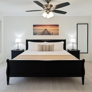 A bedroom with a black bed, two bedside tables with lamps, a ceiling fan, and a sunset painting on the wall. - Prime Location! Cozy Villa Near Disney & More (Kissimmee)