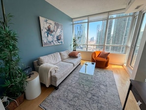 A modern living room with a sofa, a coffee table, and a view of tall buildings through large windows. - Toronto Views | 2BR | Raptors - Jays - CN Tower (Toronto)