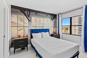 Ein modernes Schlafzimmer mit einem großen Bett, einem Schreibtisch und Blick auf eine Stadtlandschaft durch große Fenster.