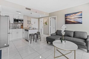 Eine moderne Küche mit weißen Schränken, einem Edelstahl-Kühlschrank und einem eingebauten Ofen. Es gibt eine Essecke mit zwei Barhocker und einem grauen Sofa. Ein runder, weißer Tisch mit goldfarbener Basis steht im Vordergrund.