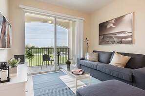 A modern living room with a grey sofa, a coffee table, and a balcony with a view of the sky and greenery. - Vista Cay 3BR Condo Near Universal & Occc (Orlando)
