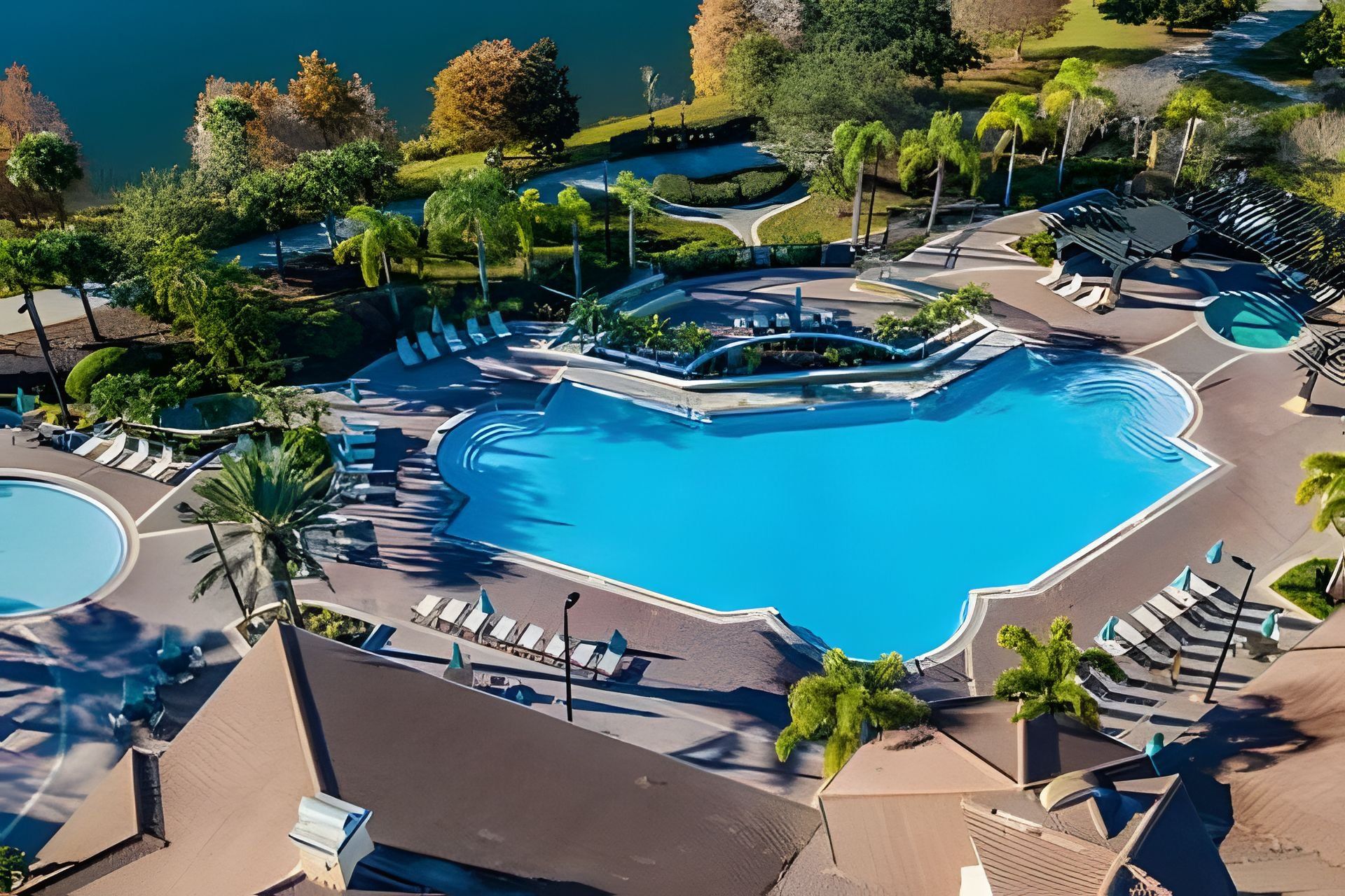 An aerial view of a resort with a large central pool, surrounded by lounge areas and palm trees.