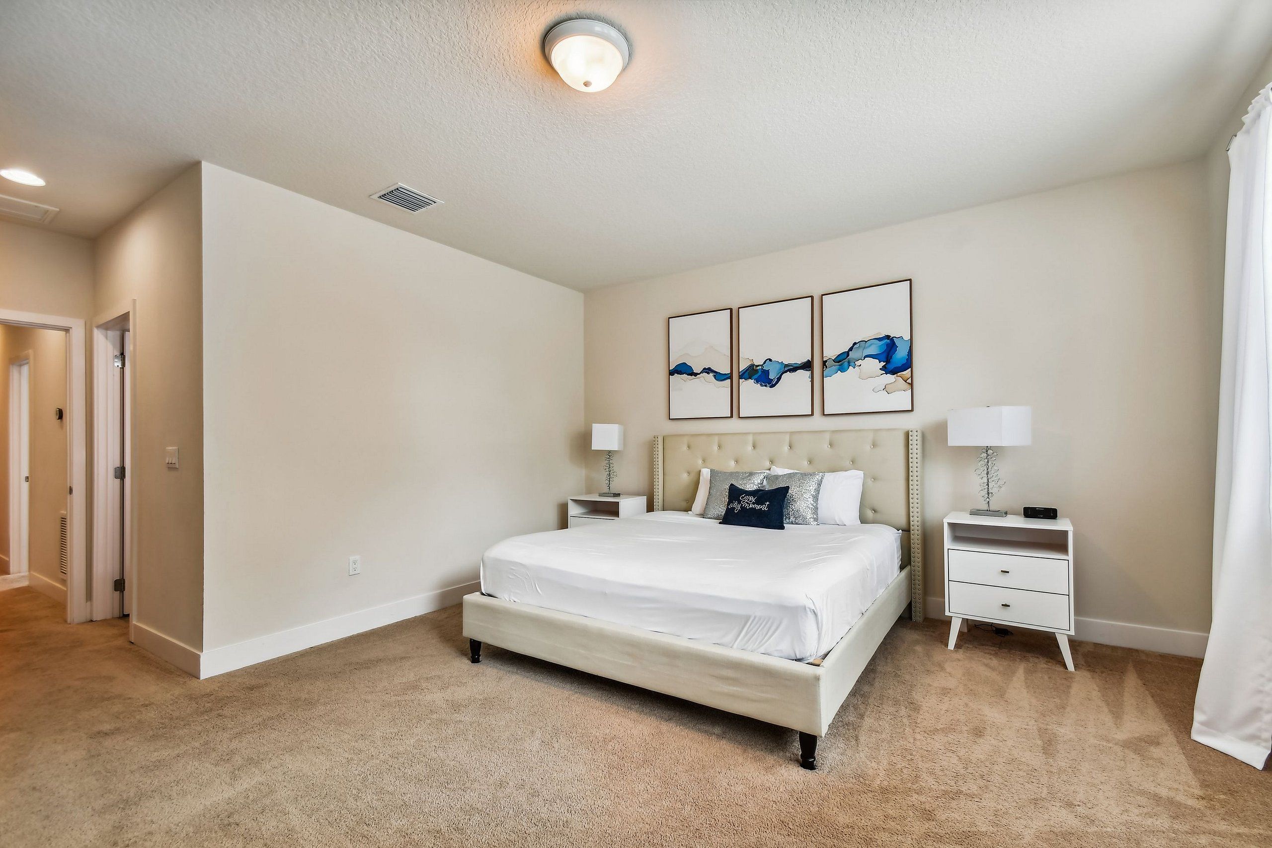 A bedroom with a large bed, a nightstand, a wall-mounted lamp, and three framed artworks.