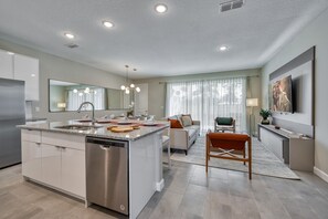 A modern kitchen with a dishwasher, a dining area with a table and chairs, and a living room with a sofa and a TV. - 4BR Villa w/ Pool Near Disney + Themed Rooms (Kissimmee)