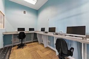 Een kamer met vier bureaus, elk met een computermonitor, toetsenbord en muis. Er zijn twee zwarte bureaustoelen. De muren zijn lichtblauw en de vloer is van hout.