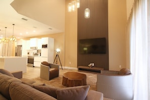 A modern living room with a large flat-screen TV, a dining area with a kitchen in the background, and a cozy seating area with sofas and a coffee table. - Modern 4 Suites Home Magic Village (Kissimmee)
