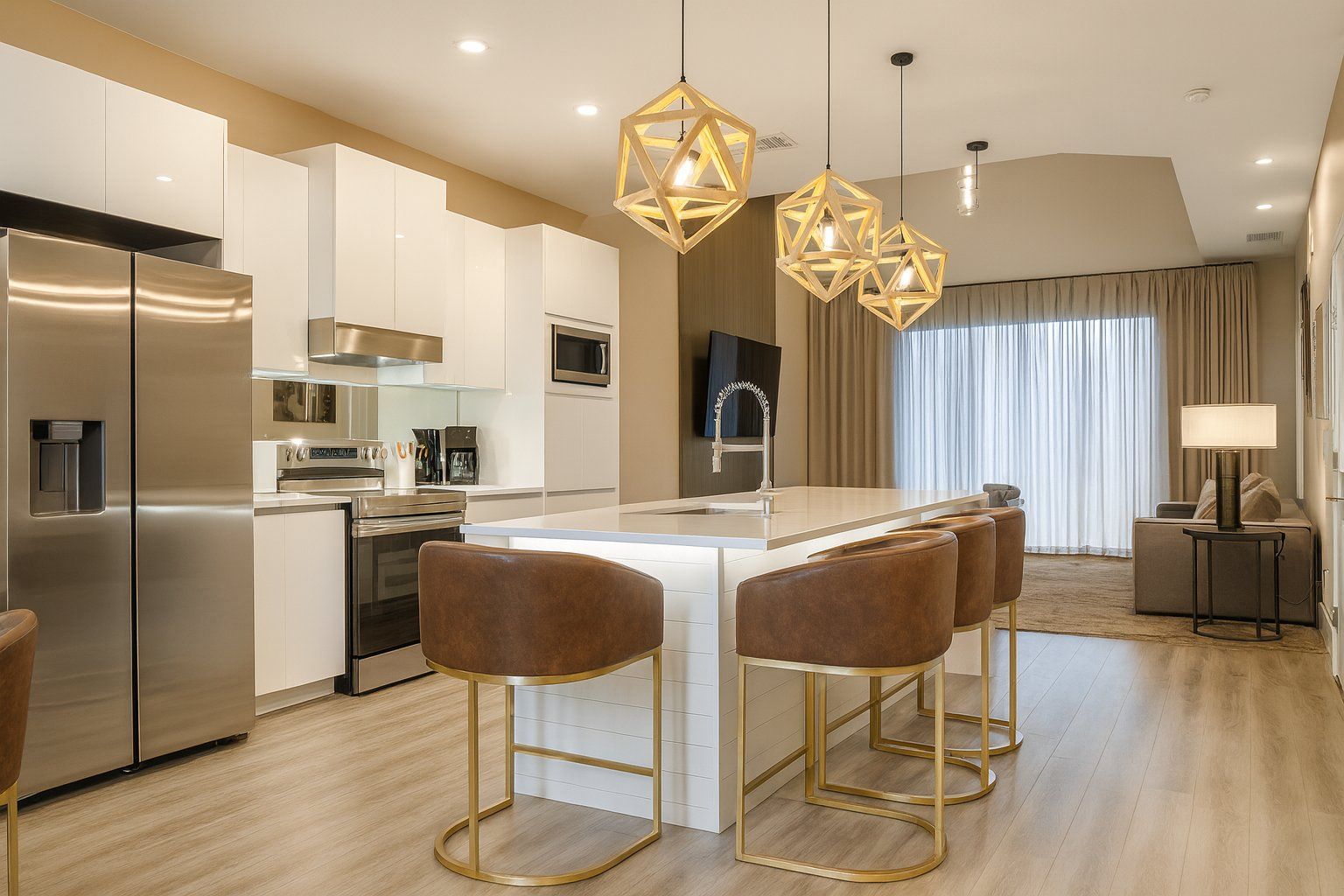 Una cocina moderna con electrodomésticos de acero inoxidable, una isla central con taburetes de bar, iluminación colgante y una sala de estar con sofá y lámpara.