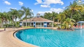 A swimming pool with a clear blue water, surrounded by palm trees and a building with a blue roof. - 3BR Lucaya Village Home Near Disney (Kissimmee)