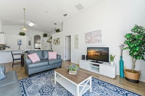 A modern living room with a grey sofa, a coffee table, and a TV. There is a kitchen in the background with white cabinets and a dining area. - Single-story 4BR Home w/ Pool & Game Room (Kissimmee)