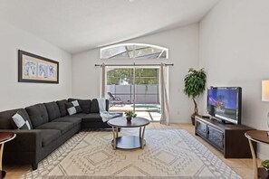 A modern living room with a sectional sofa, a round coffee table, a flat-screen TV, and a large window with curtains. - 4BR Home w/ Private Pool & Spa Near Disney (Kissimmee)