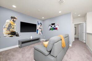 A modern living room with a grey sofa, a flat-screen TV, and sports-themed wall art. - Casa 3Q c/ Piscina e Game Room Disney (Clermont)
