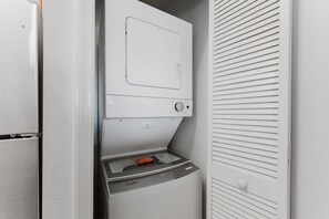 A compact laundry room with a front-loading washer and dryer, a small countertop, and a door with horizontal louvered panels. - Resort Suite on International Drive (Orlando)