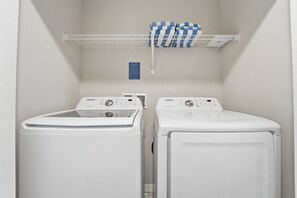 A laundry room with two white Samsung washing machines, a metal shelf with folded laundry, and a blue informational sign. - Fiesta Key 3BR Townhome Near Disney (Kissimmee)