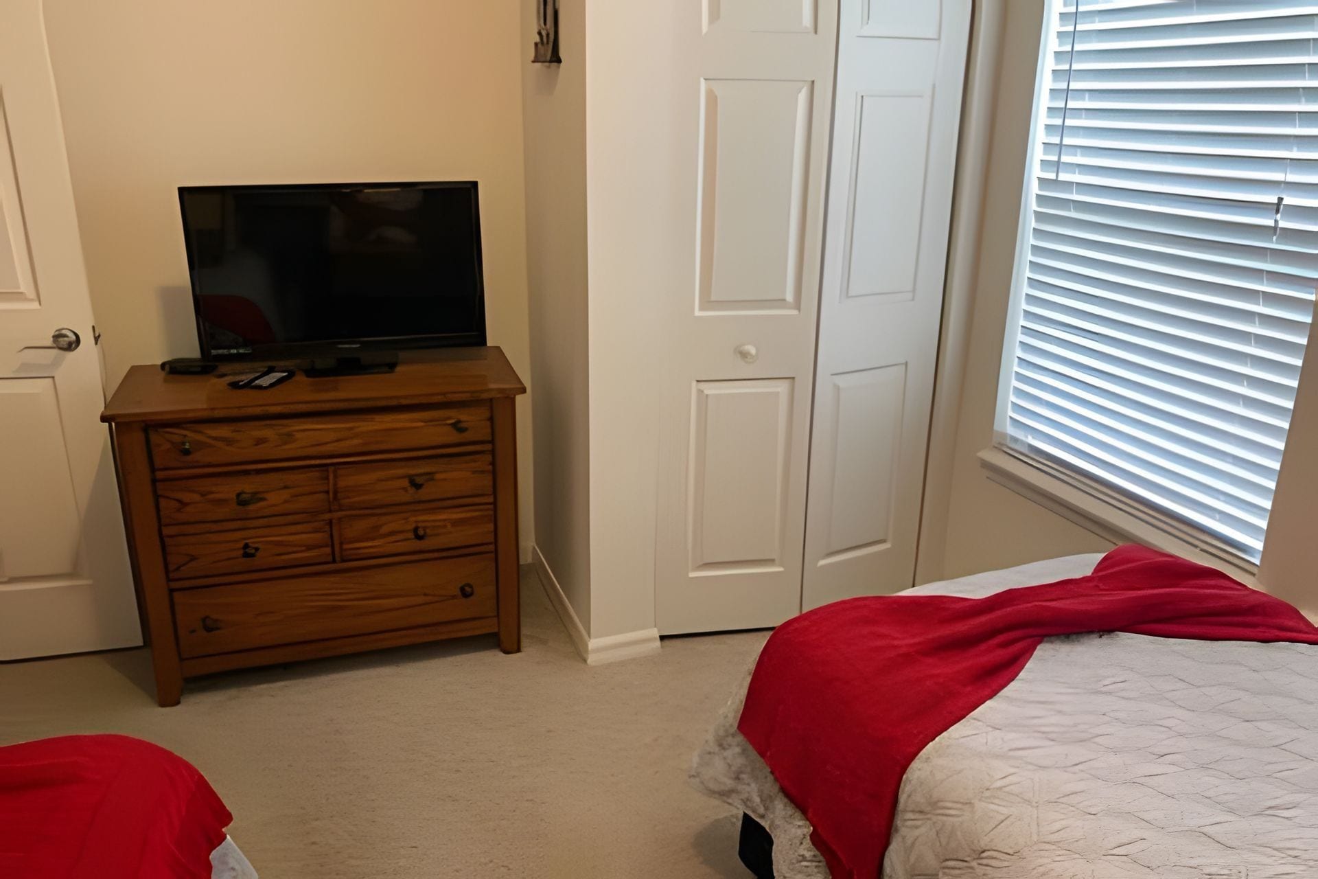 Ein Schlafzimmer mit einem Holzkapuzenpaneel, einem Fernseher darauf, einem Bett mit roter Decke und einem Fenster mit Jalousien.