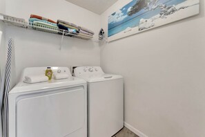A laundry room with two white front-loading washers and dryers, a shelf with folded towels, and a painting of a beach scene on the wall. - 5BR Lake Berkley Home, Lake-view Pool (Kissimmee)