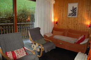 A cozy room with a wooden wall, a sofa, two armchairs, and a view of greenery outside.
