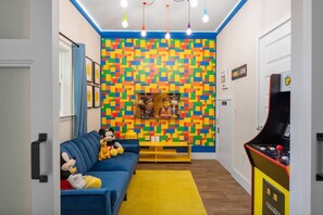 A room with a colorful LEGO wall, a blue sofa with plush toys, a yellow rug, and a retro arcade machine. - Renovated 4BR Townhome w/ Disney Game Room (Kissimmee)
