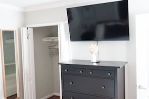A room with a flat-screen TV mounted above a dark-colored dresser with drawers, a white vase with dried flowers, and a wooden door leading to a closet with a hanging rack. - Bright DC Retreat / 3BR / Free Parking (Washington)