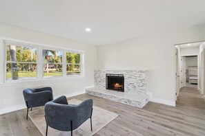 A modern living room with a fireplace, two grey armchairs, and a large window overlooking a garden. - 4BR Lakefront Home w/ Dock & Private Views (Orlando)