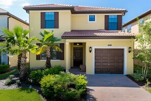 A two-story house with a brown garage door, a small porch, and a well-maintained garden with a palm tree and shrubs. - Solterra Villa De Tommaso 5BR w Pool & Games (Davenport)