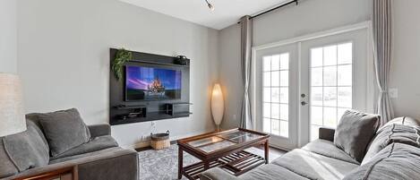 A modern living room with a grey sofa, a coffee table, a TV mounted on the wall, and a large window with curtains.