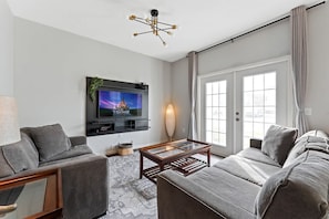 Ein modernes Wohnzimmer mit einem grauen Sofa, einem Couchtisch, einem an der Wand montierten Fernseher und einem großen Fenster mit Vorhängen.