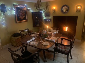 A dining area with a wooden table set for four, surrounded by chairs, a fireplace, and wall-mounted lights. - Cottage Sleeps 7 With Garden & Free Parking (Dorchester)