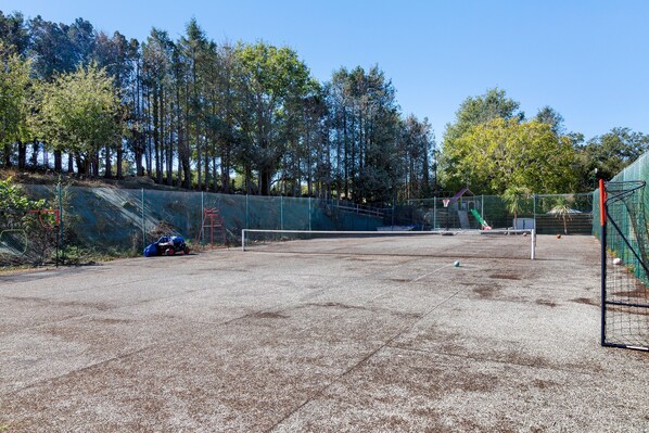 Una cancha de tenis exterior con red, rodeada por una valla y árboles.
