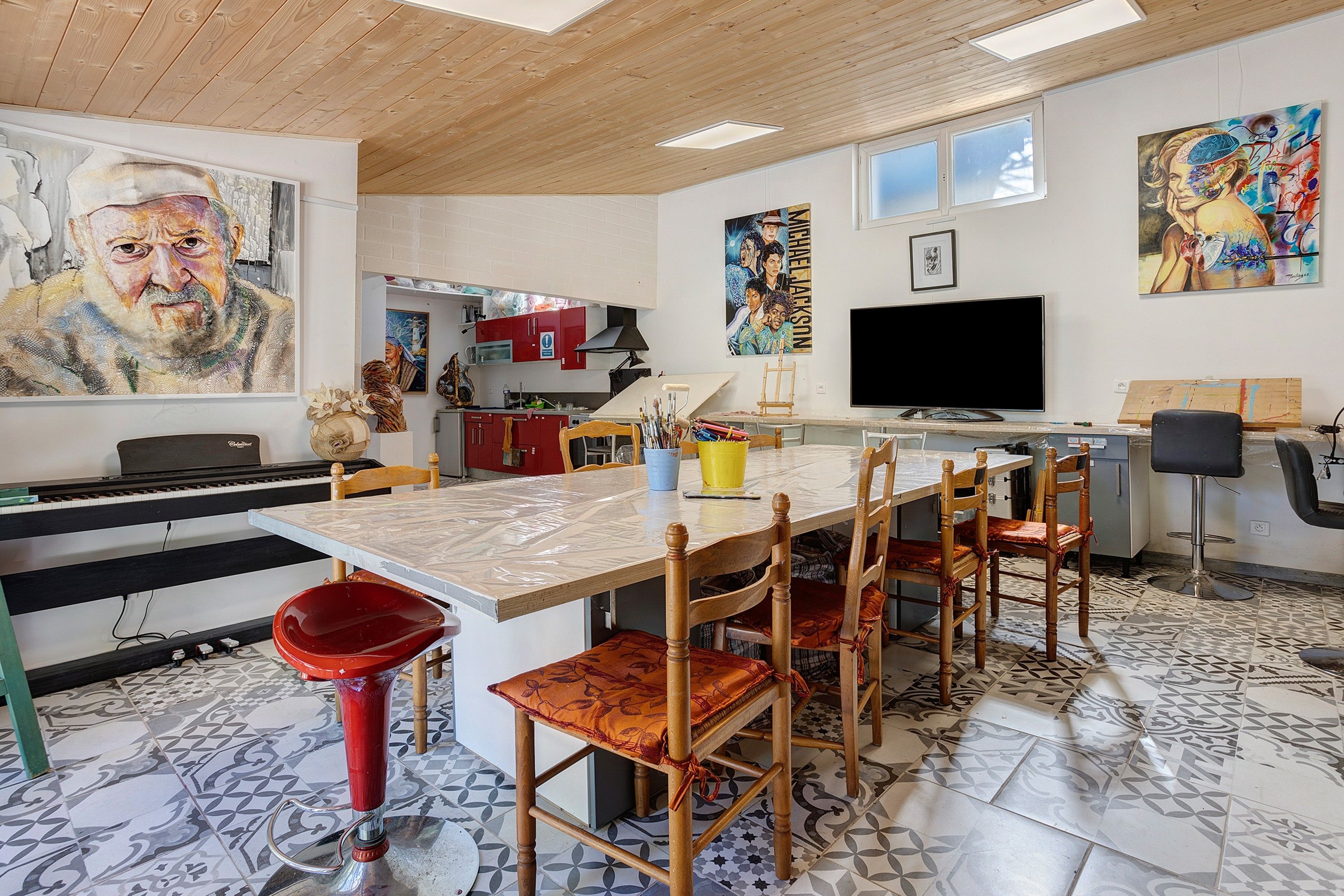 A modern kitchen with a dining area, featuring a large painting, a television, and a piano.