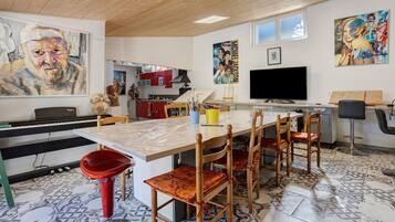 A modern kitchen with a dining area, featuring a large painting, a television, and a piano.