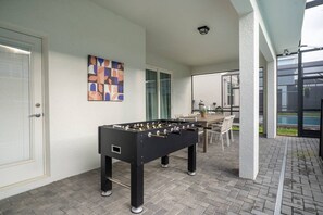 A foosball table, a dining table, and chairs in an outdoor patio area with a view of a swimming pool. - 6BR Disney Home w/ Pool at Windsor Cay (Clermont)
