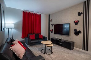 A modern living room with a black sofa, red curtains, a flat-screen TV, and Mickey Mouse wall decorations. - 6BR Villa w Pool & Game Room Near Disney (Davenport)