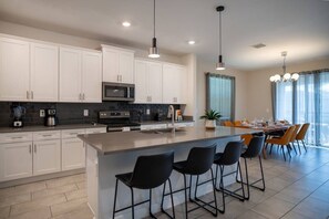 A modern kitchen with white cabinets, a central island with bar stools, a dining area with a table and chairs, and a large window with curtains. - 6BR Villa w Pool & Game Room Near Disney (Davenport)