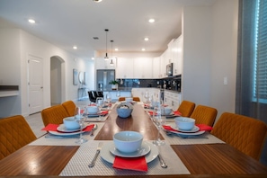 A modern dining room with a long wooden table set for six, surrounded by orange chairs, and a kitchen in the background. - 6BR Villa w Pool & Game Room Near Disney (Davenport)