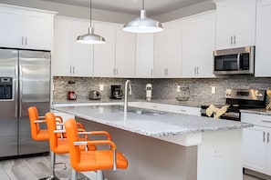 A modern kitchen with white cabinets, a large island with orange bar stools, stainless steel appliances, and pendant lighting. - 6BR Townhome w/ Pool 10 Min to Disney (Kissimmee)