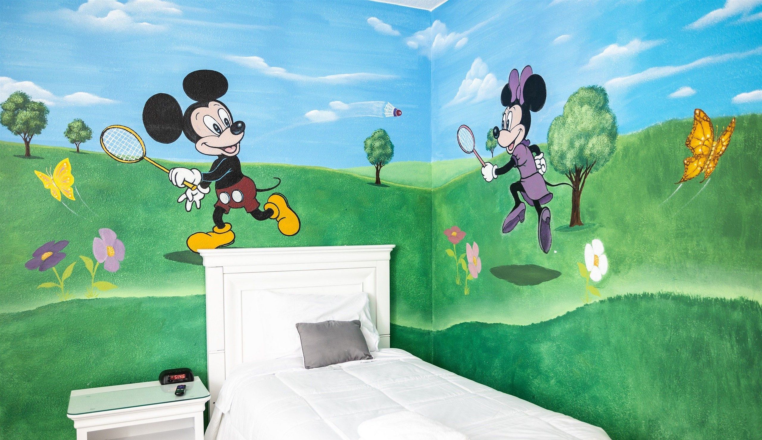 A bedroom with a bed, a nightstand, and a wall mural featuring cartoon characters playing tennis.