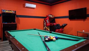 Ein Billardtisch mit Kugeln und Spielstangen, ein rotes Arcade-Spielgerät und ein Fernseher an der Wand.