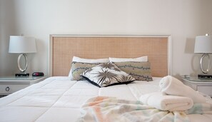 Ein ordentlich bezogenes Bett mit weißer Decke und zwei Zierkissen, ein Nachttisch mit Lampe und einer digitalen Uhr.