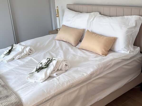A neatly made bed with white and beige bedding, a bedside table with a lamp, and a wall-mounted light fixture.
