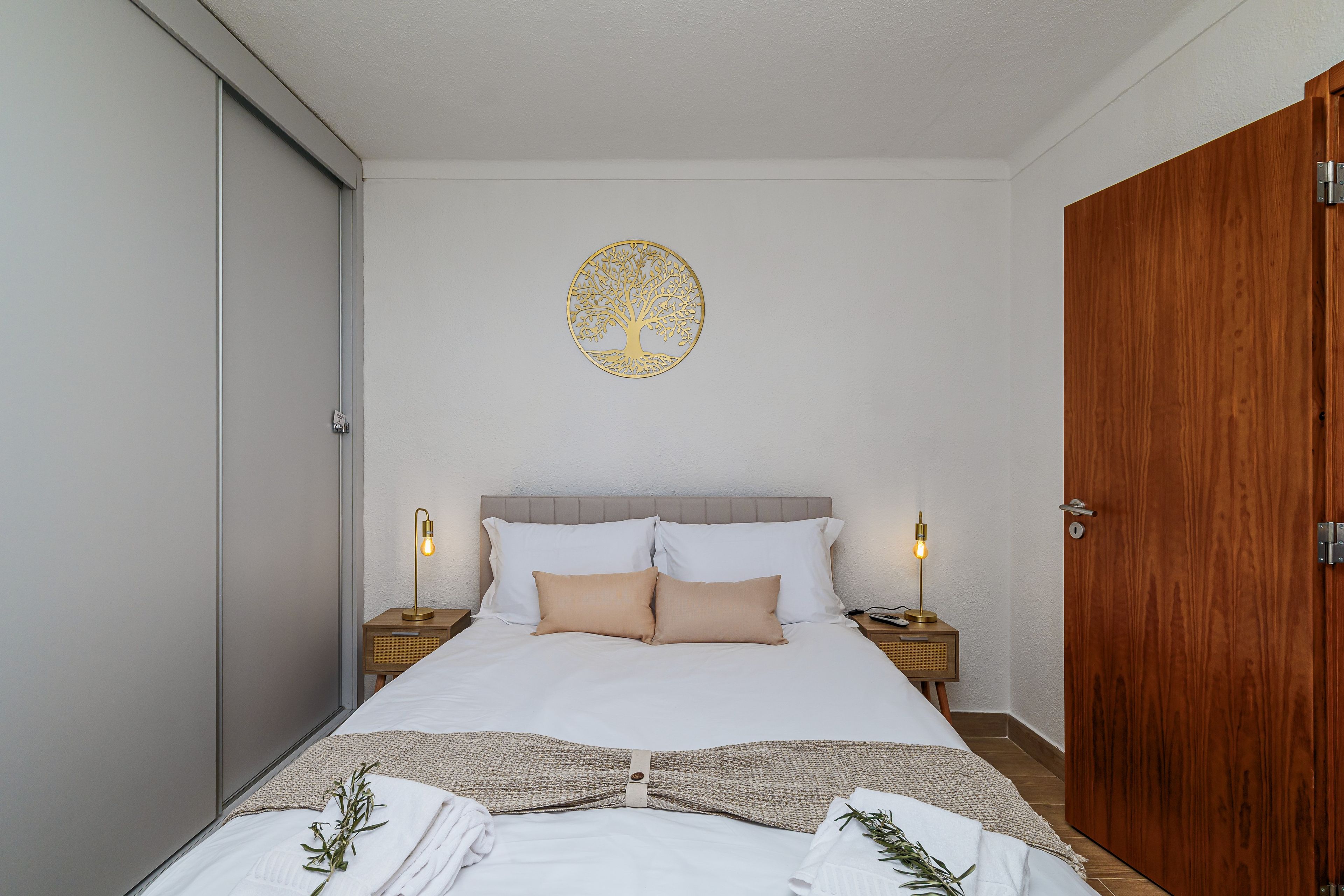 Una cama bien hecha con sábanas blancas y dos almohadas, flanqueada por mesitas de noche con lámparas, en una habitación con puerta de madera y un adorno en la pared.