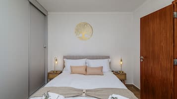 Una cama bien hecha con sábanas blancas y dos almohadas, flanqueada por mesitas de noche con lámparas, en una habitación con puerta de madera y un adorno en la pared.