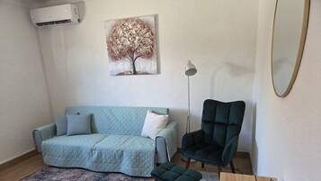 A room with a light blue sofa, a dark armchair, a small stool, a painting of a tree, a floor lamp, and a wooden table with a vase.